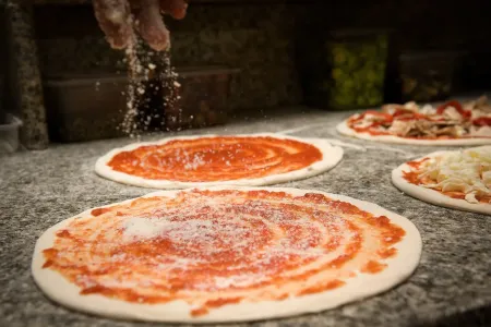 Pertsona batek parmesano gazta botatzen dio saltsarekin pizzari mostradorean, beste pizza batzuk atzealdean daudela.