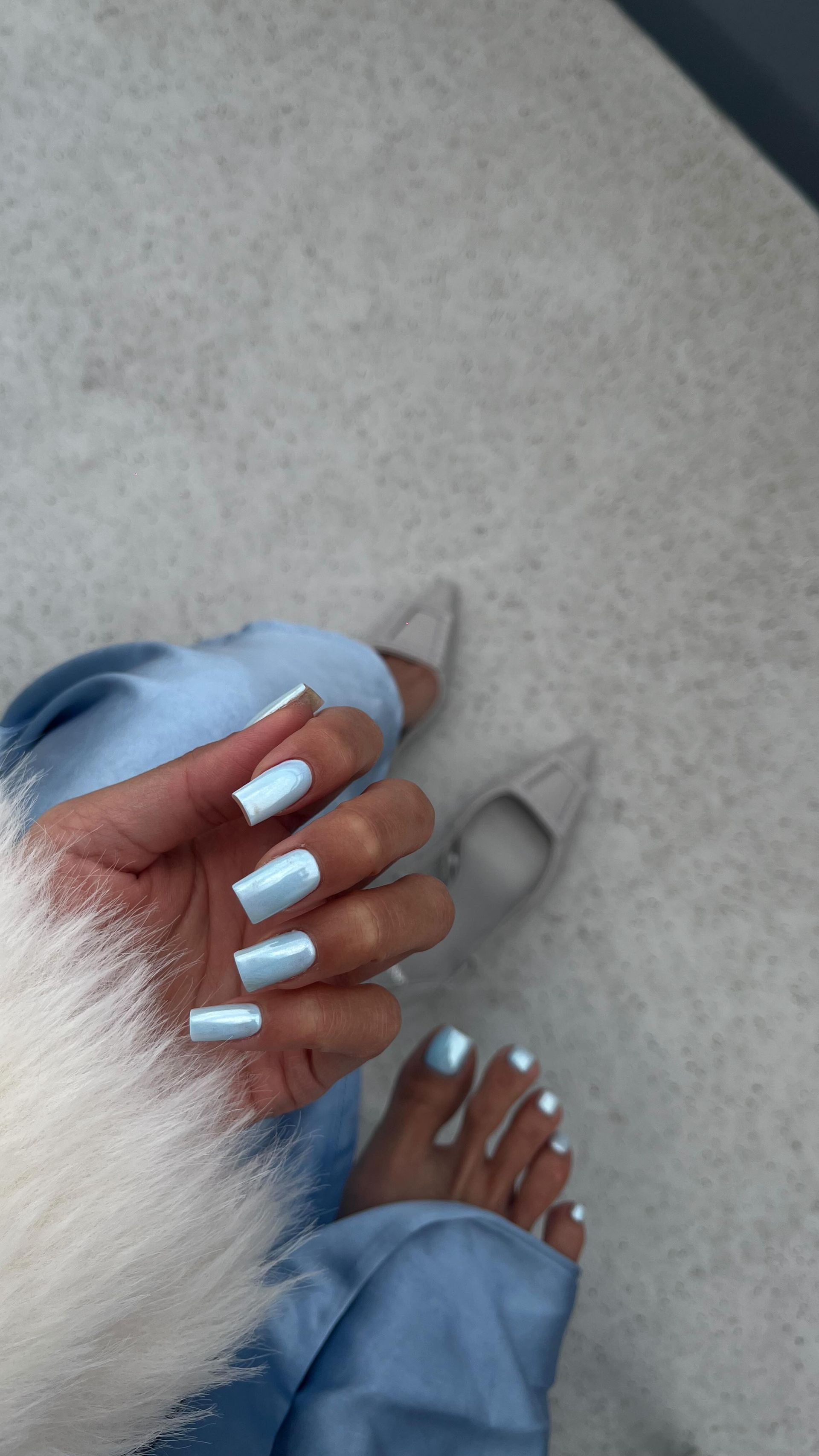 Hände mit pastellblauen Nägeln und flauschigen weissen Ärmeln über hellblauen Jeans und weissen Schuhen auf einem grauen Boden