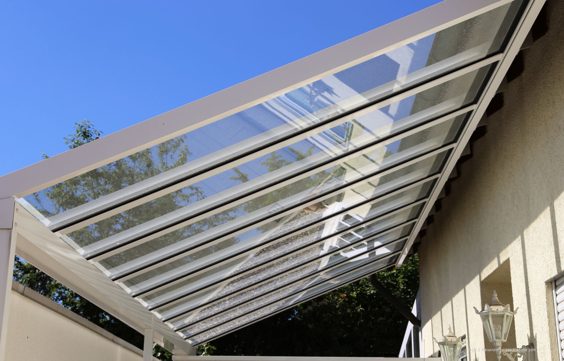 Eine weiße Lamellenmarkise überspannt eine Terrasse neben einer beigen Mauer; durch die Struktur sind blauer Himmel und Bäume sichtbar.