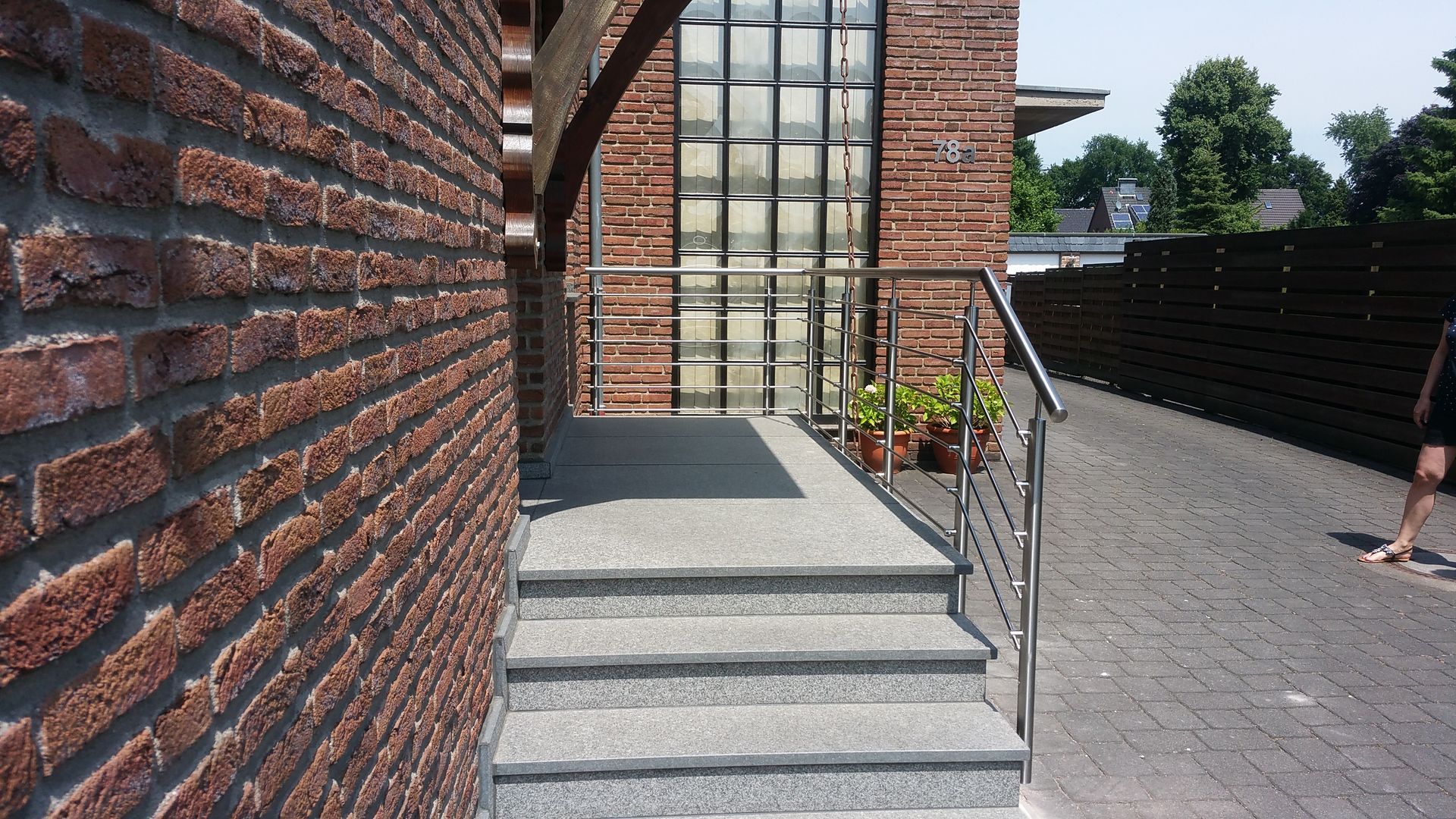 Eingang zum Backsteinhaus mit grauen Stufen, Metallgeländer und einer Glastür, die zu einem Kiesweg führt.
