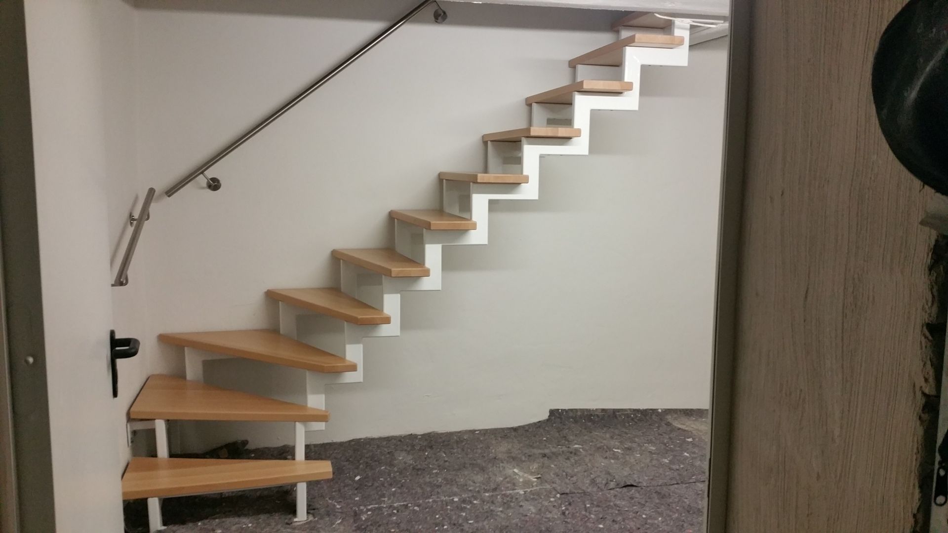 Freitragende Holztreppe mit offenen Setzstufen vor einer weißen Wand in einem unfertigen Innenraum.
