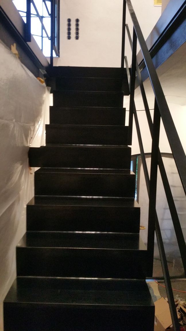 Eine schmale, schwarze Treppe mit Metallgeländer führt zwischen zwei Wänden nach oben.
