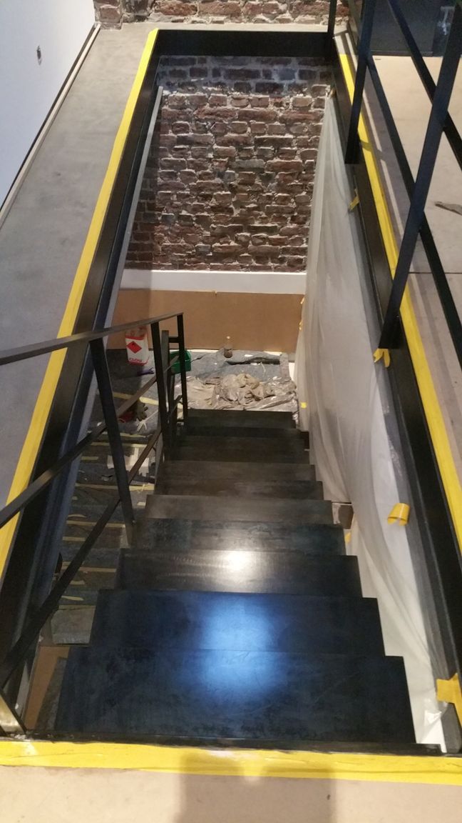 Steile Innentreppe mit schwarzen Stufen, gelben Sicherheitskanten und einer Backsteinmauer am Fuß.