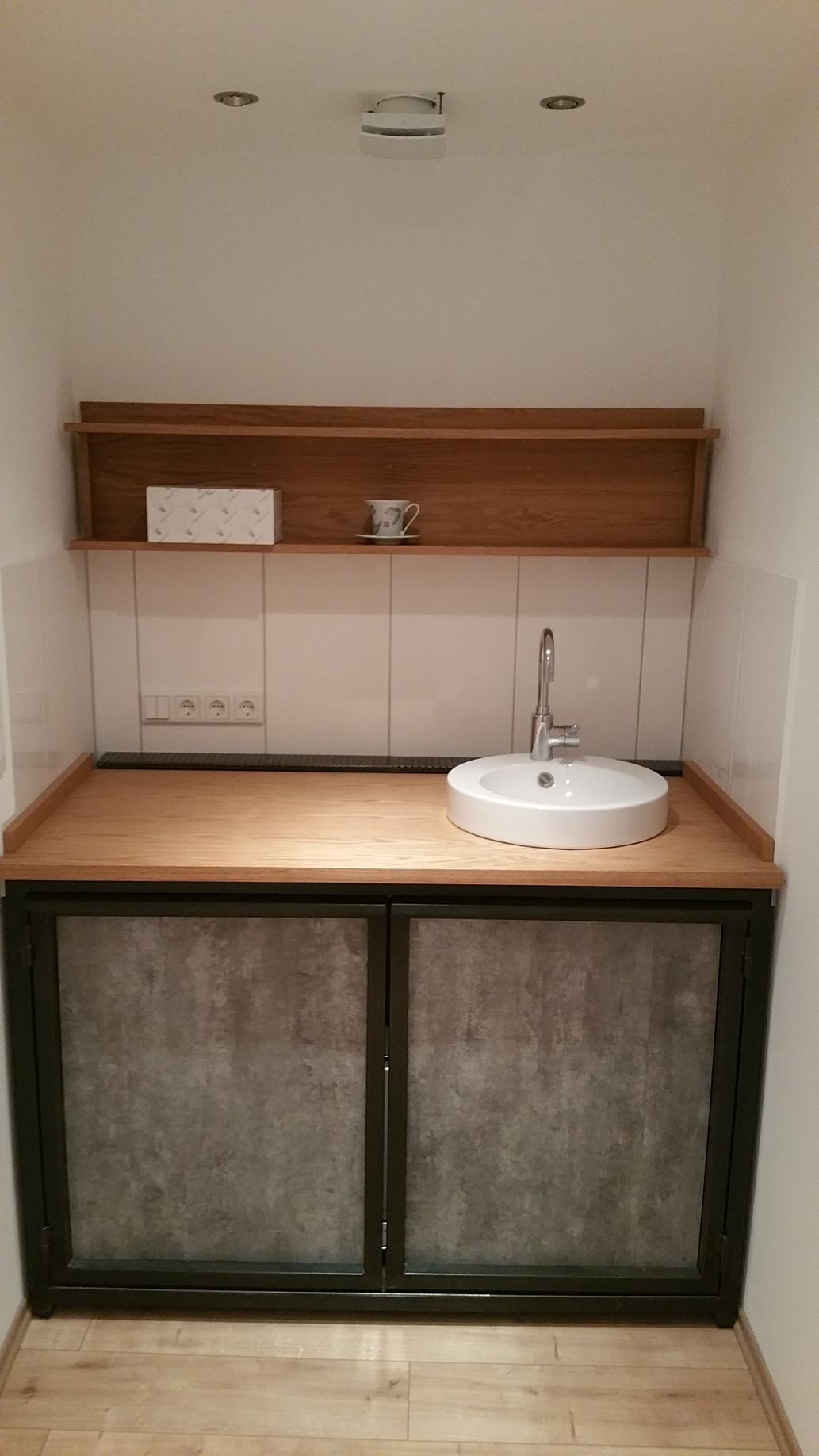Kleiner Waschtisch im Badezimmer mit Holzregalen, gefliestem Spritzschutz und einem weißen runden Waschbecken über dunklen Schranktüren.