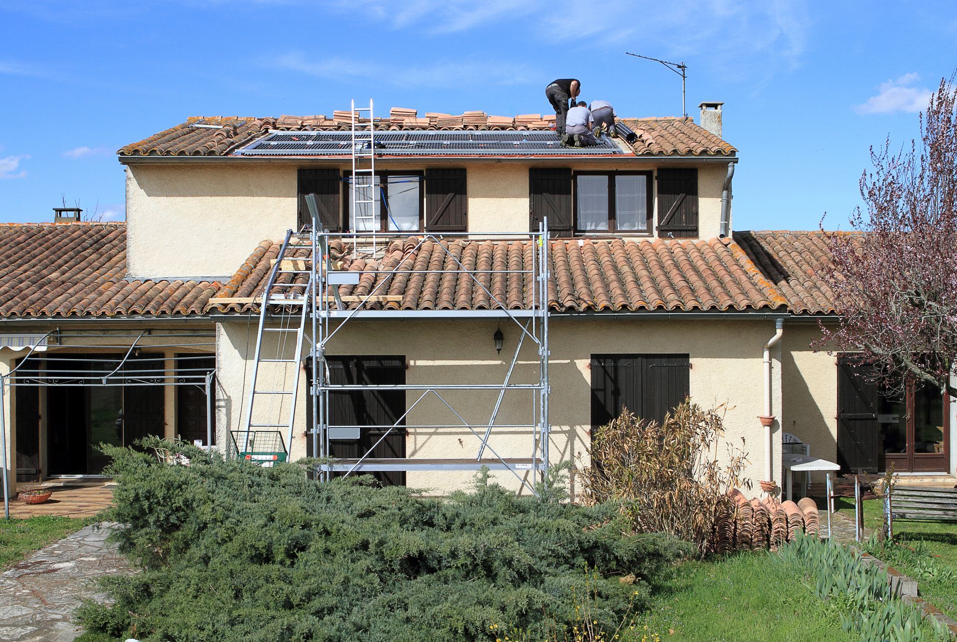 Maison avec panneaux solaires sur le toit, échafaudages et toit de tuiles brunes par une journée ensoleillée.