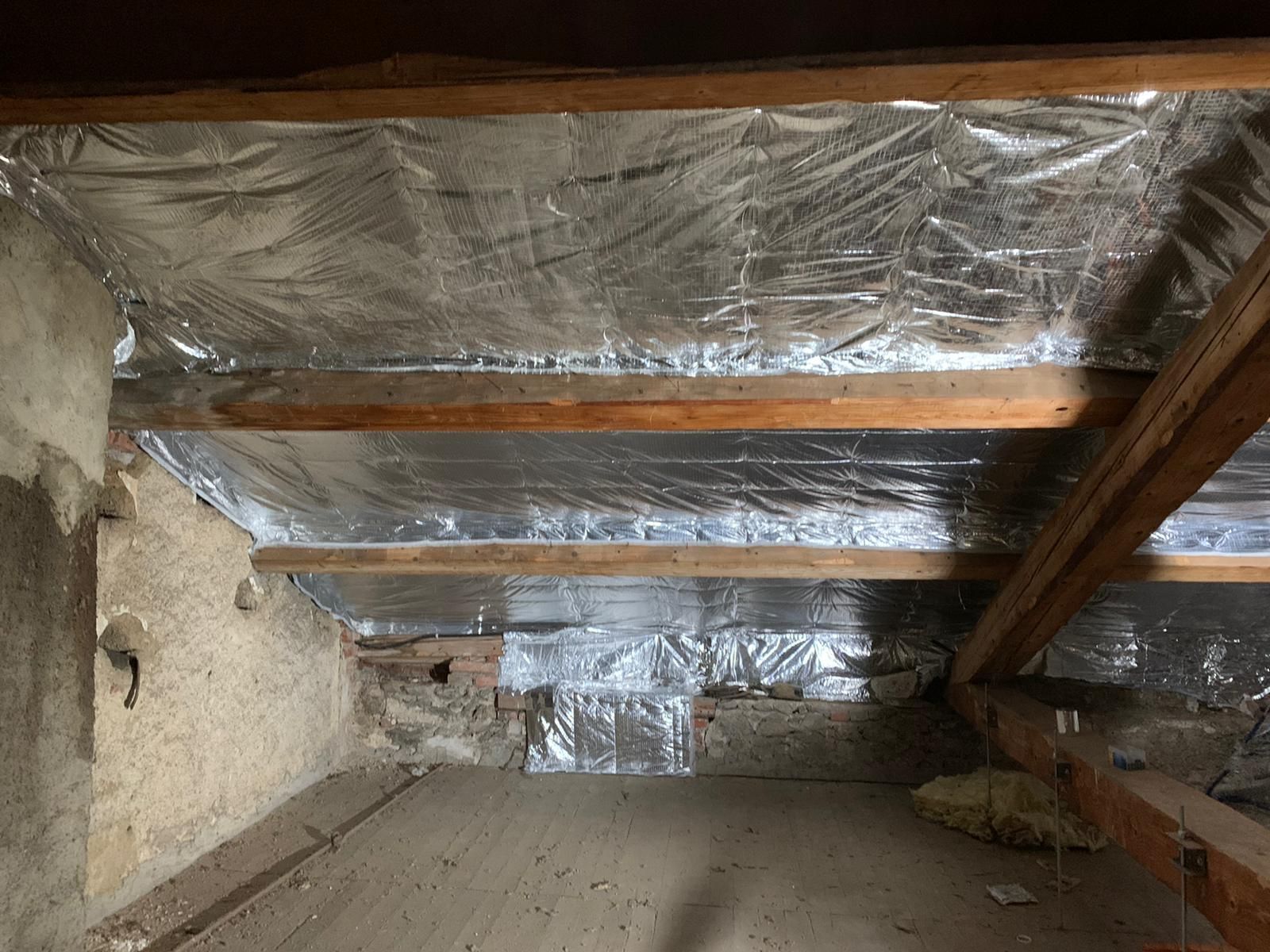 Combles aménagés avec isolation réfléchissante argentée installée entre les poutres en bois, au-dessus d'un mur en pierre apparente.