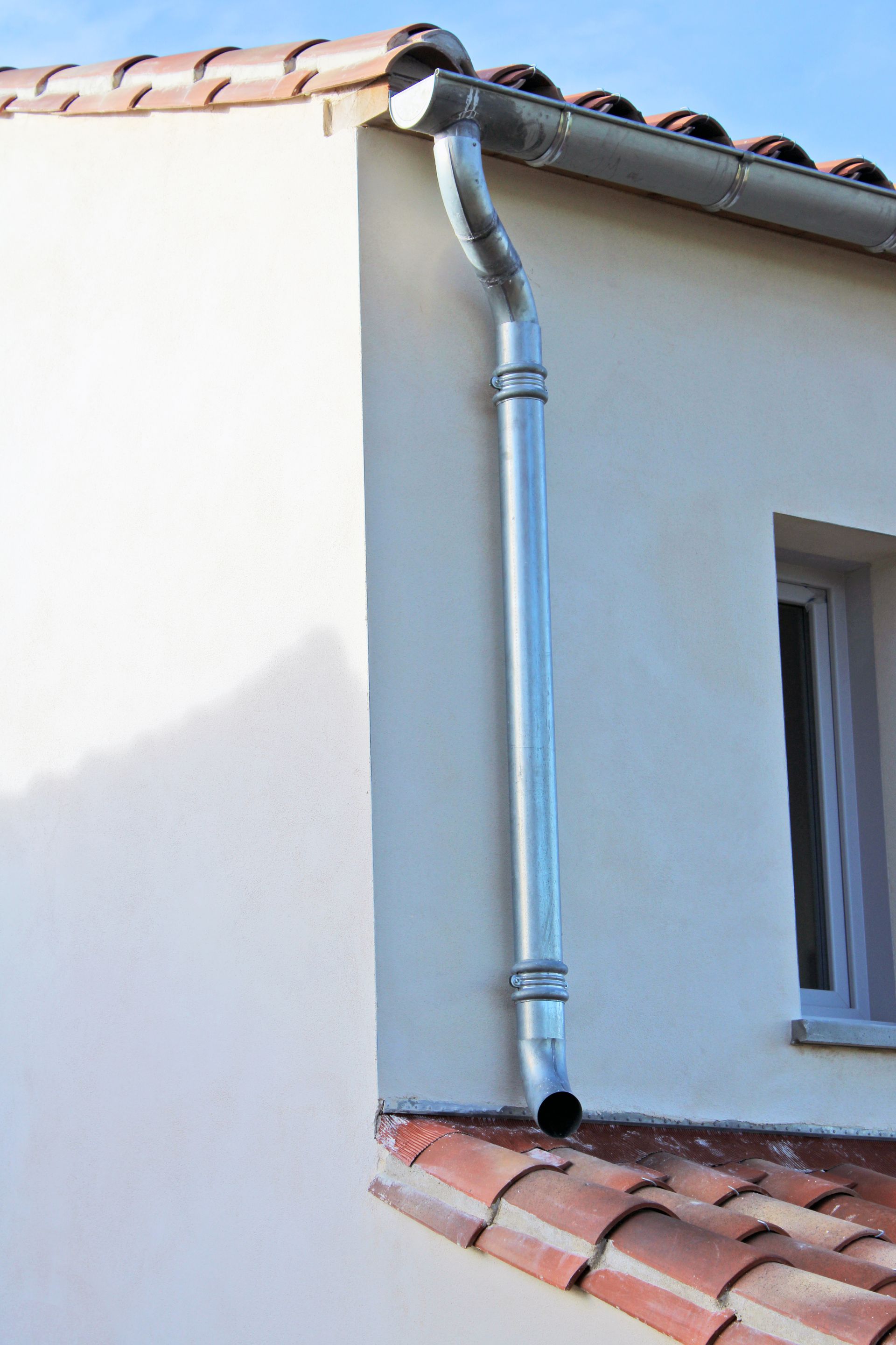 Extérieur de maison avec une gouttière et un tuyau de descente argentés sur un mur blanc, à côté d'un toit de tuiles rouges