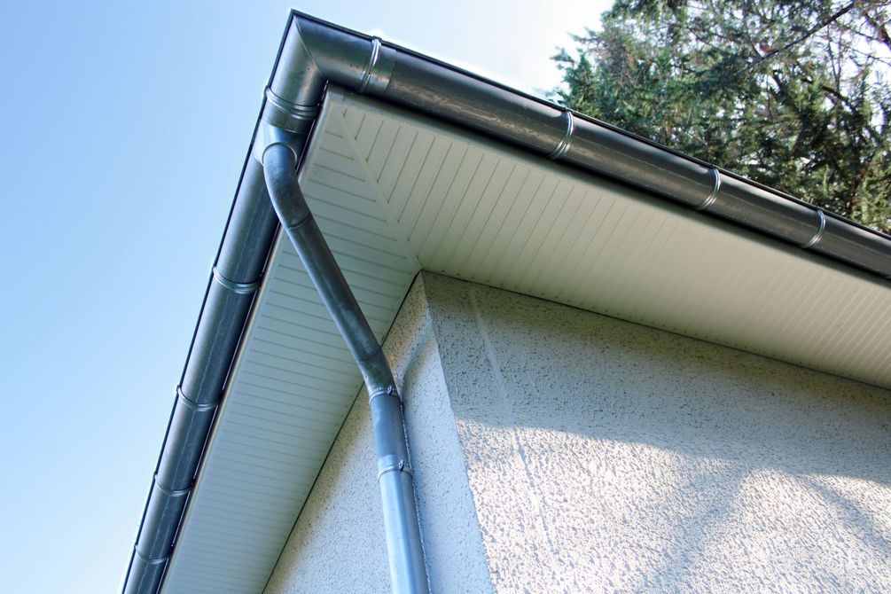 Coin d'une maison avec bardage blanc, gouttière et descente pluviale se détachant sur un ciel bleu