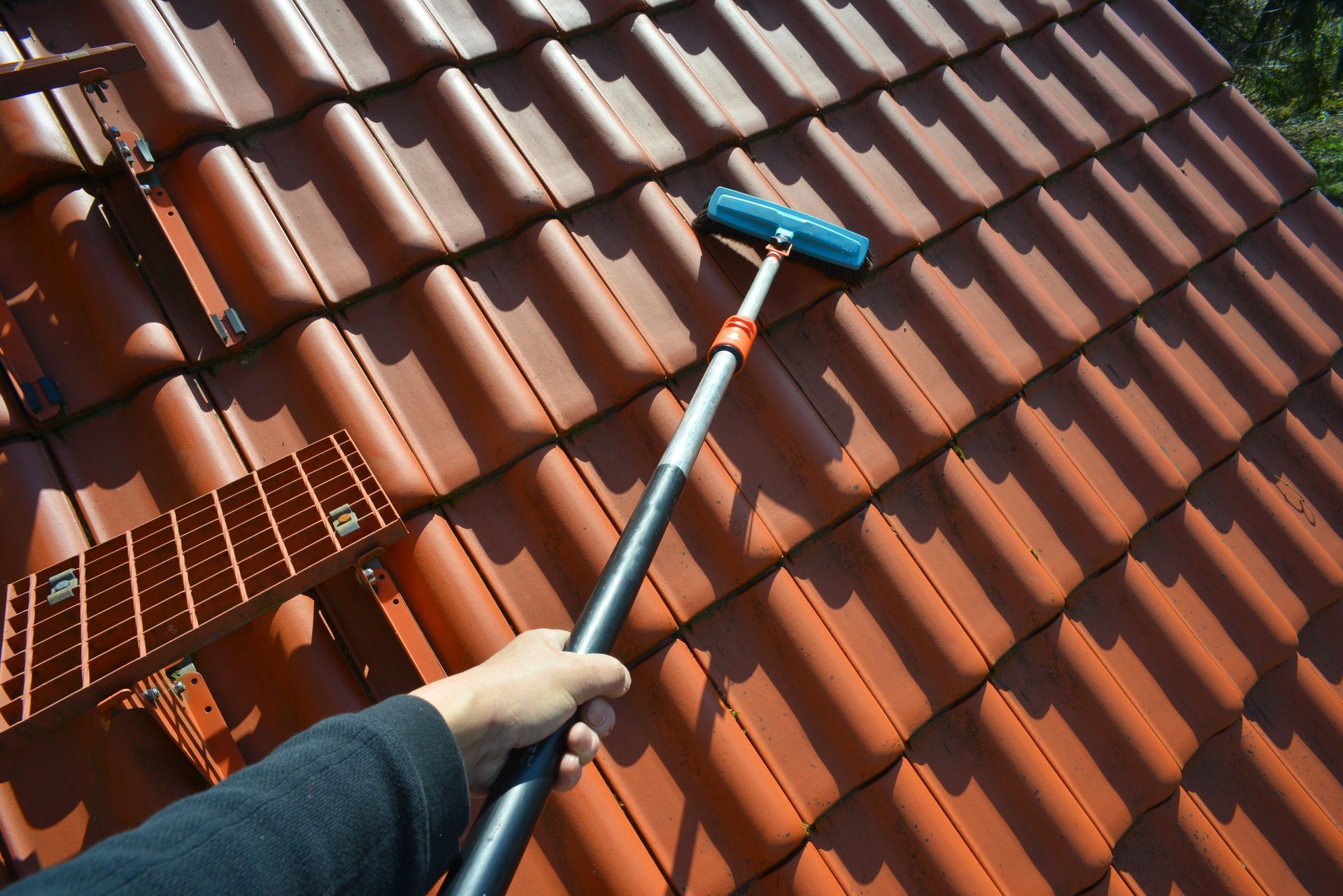 Une personne nettoie un toit de tuiles rouges avec une brosse à manche bleu fixée à une longue perche.