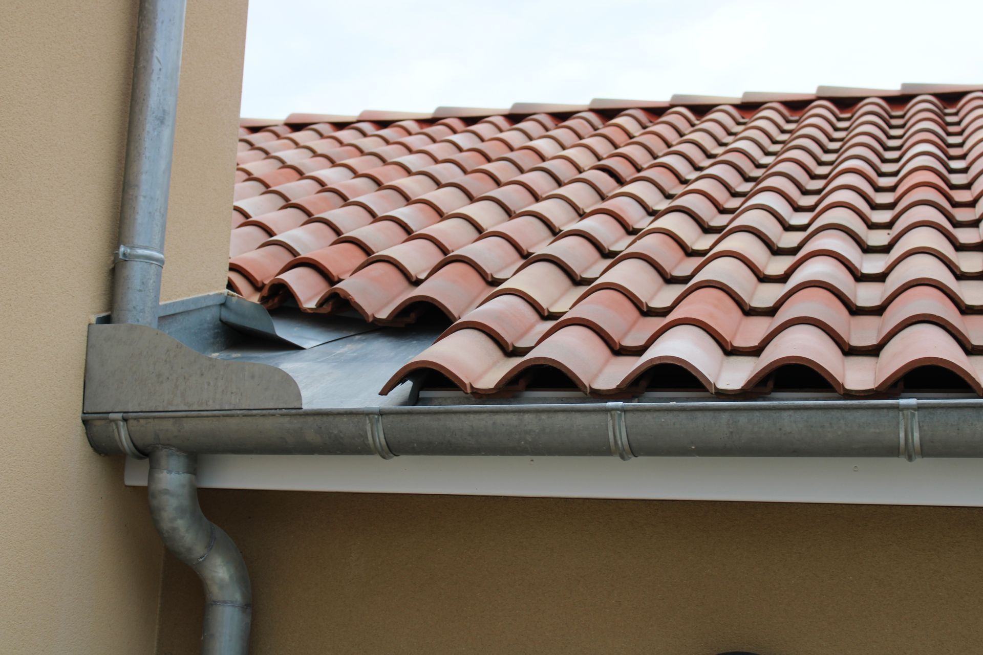 Bord de toit avec tuiles en terre cuite, gouttière et descente pluviale contre un mur beige