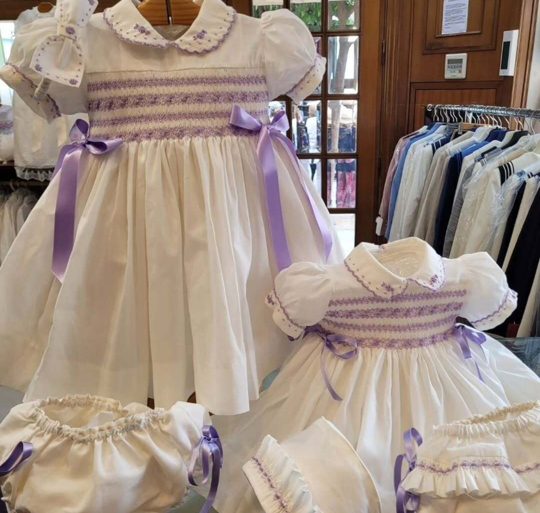 Vestidos blancos con bordados morados, lazos y mangas abullonadas; expuestos en una tienda.