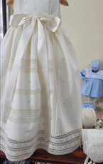Vestido de bautizo color marfil con ribete de encaje y lazo, colgante con capota azul en el fondo.