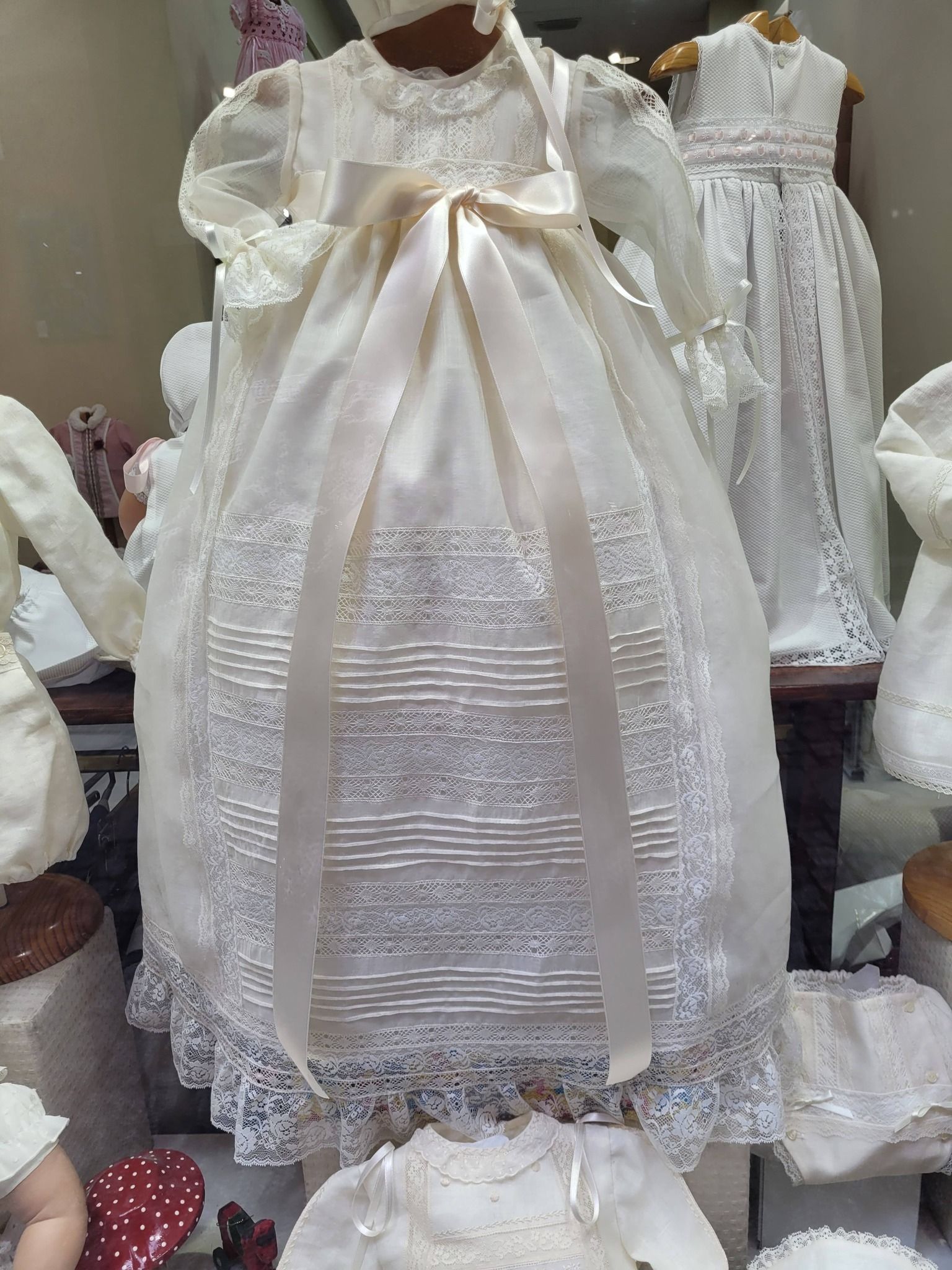 Vestido de bautizo color crema con detalles de encaje y fajín de cinta. 