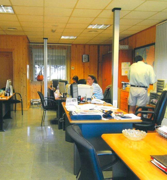 Oficina con gente trabajando en escritorios, paredes revestidas de madera, luces fluorescentes y monitores de ordenador.