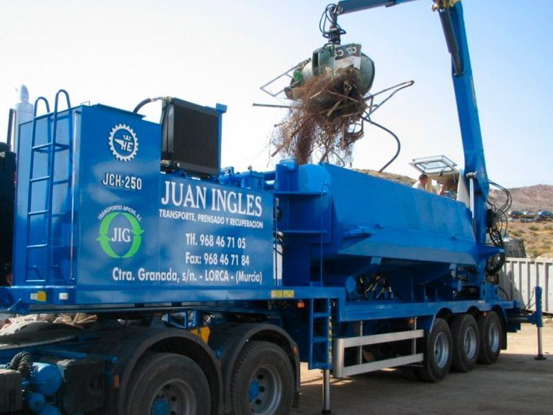 Camión azul JUAN LING con grúa descargando metal enredado en un patio de construcción.