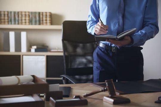 Un hombre está de pie frente a un escritorio sosteniendo un cuaderno y un bolígrafo.