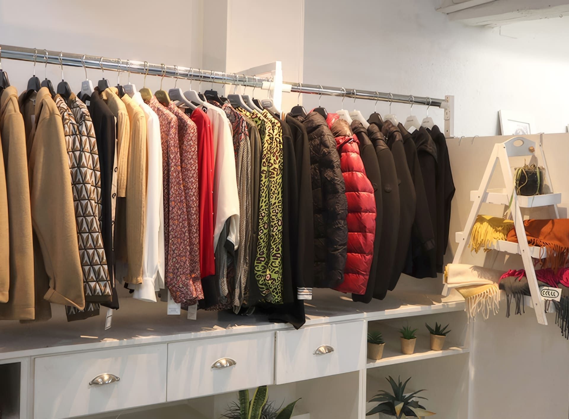 Perchero en una boutique con diversas prendas; una cómoda blanca debajo y estantes con plantas.