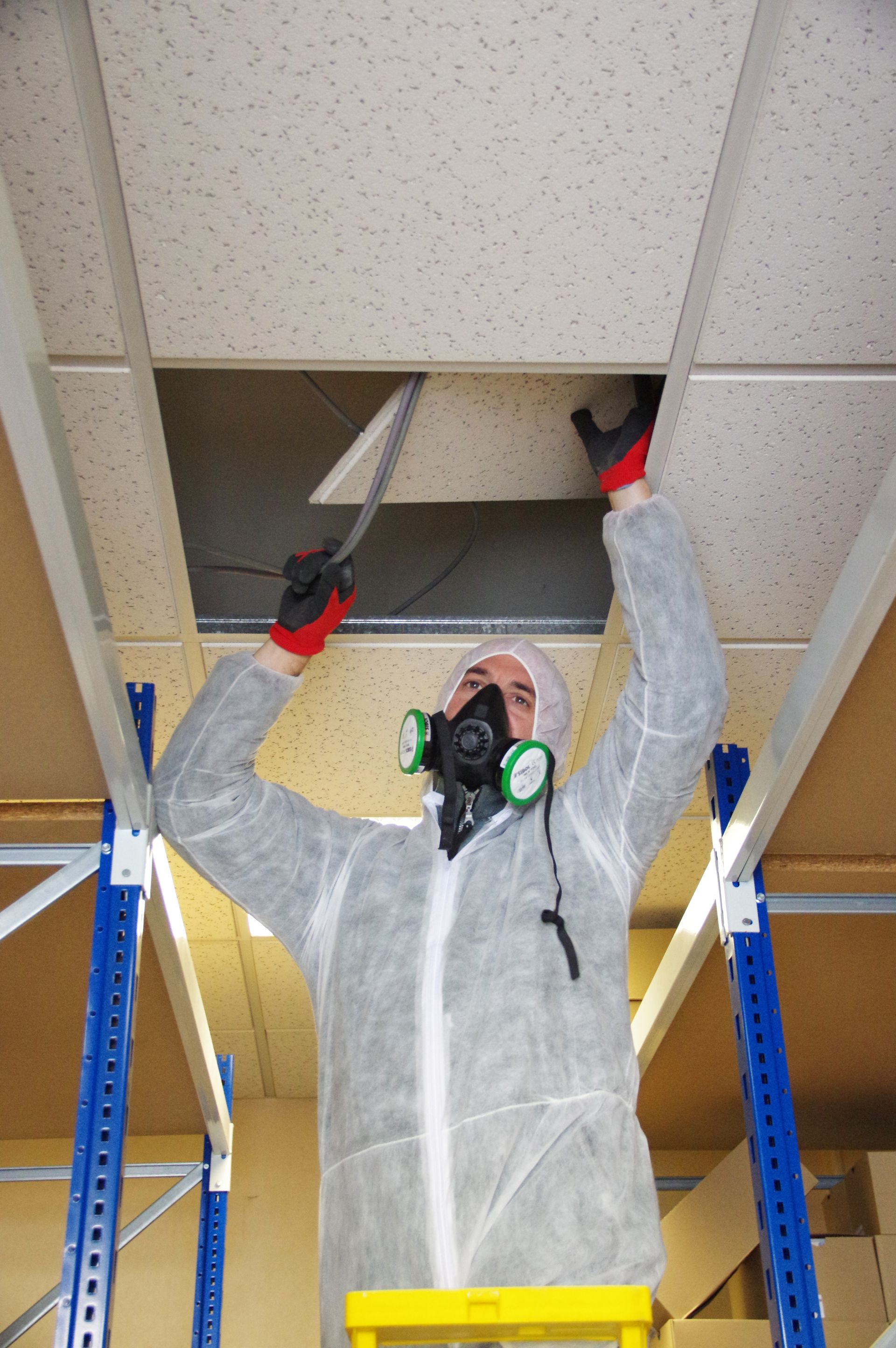 Une personne portant un équipement de protection retire une dalle de plafond. Des étagères métalliques sont visibles.