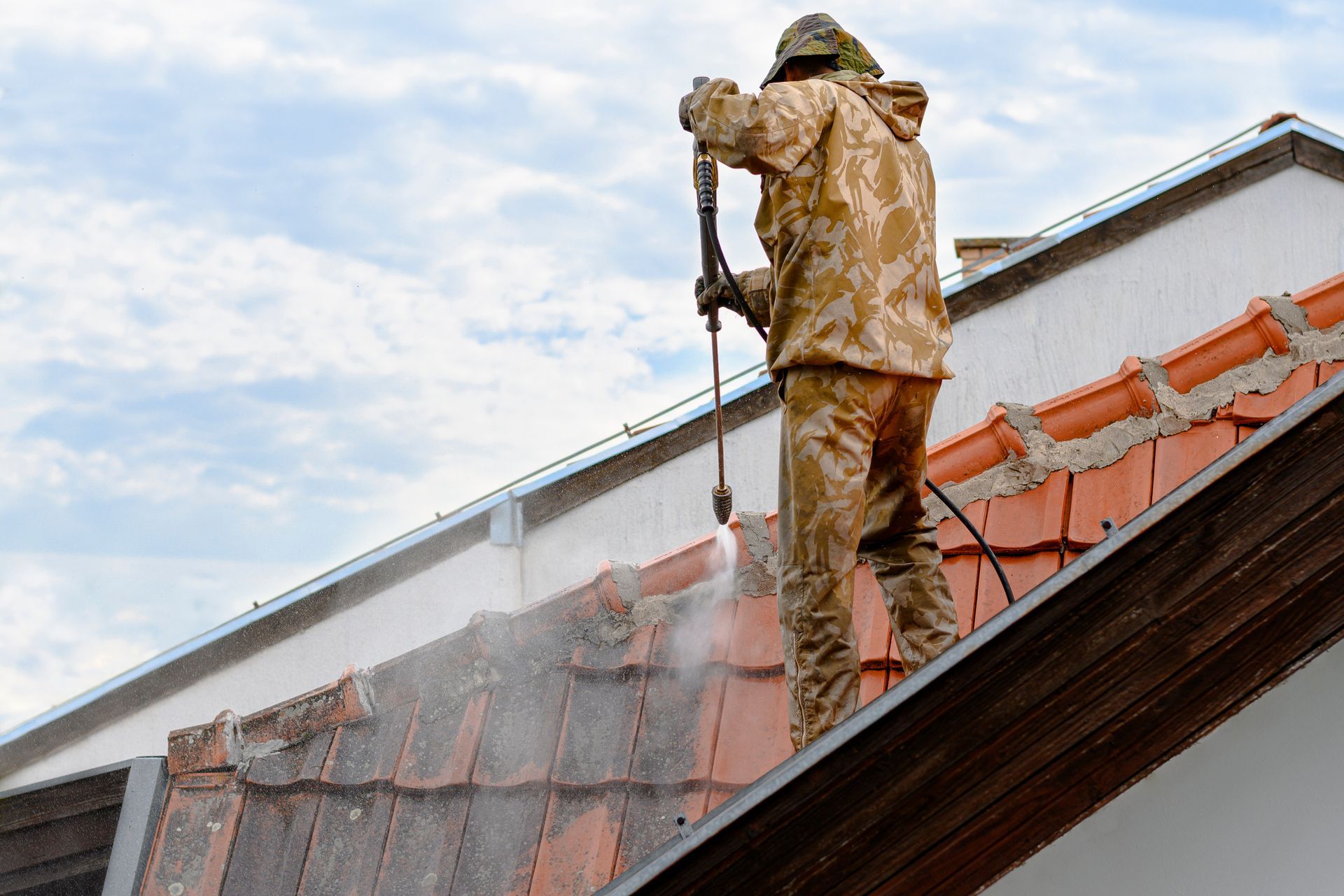 Une personne en tenue de camouflage nettoie un toit de tuiles au nettoyeur à haute pression.