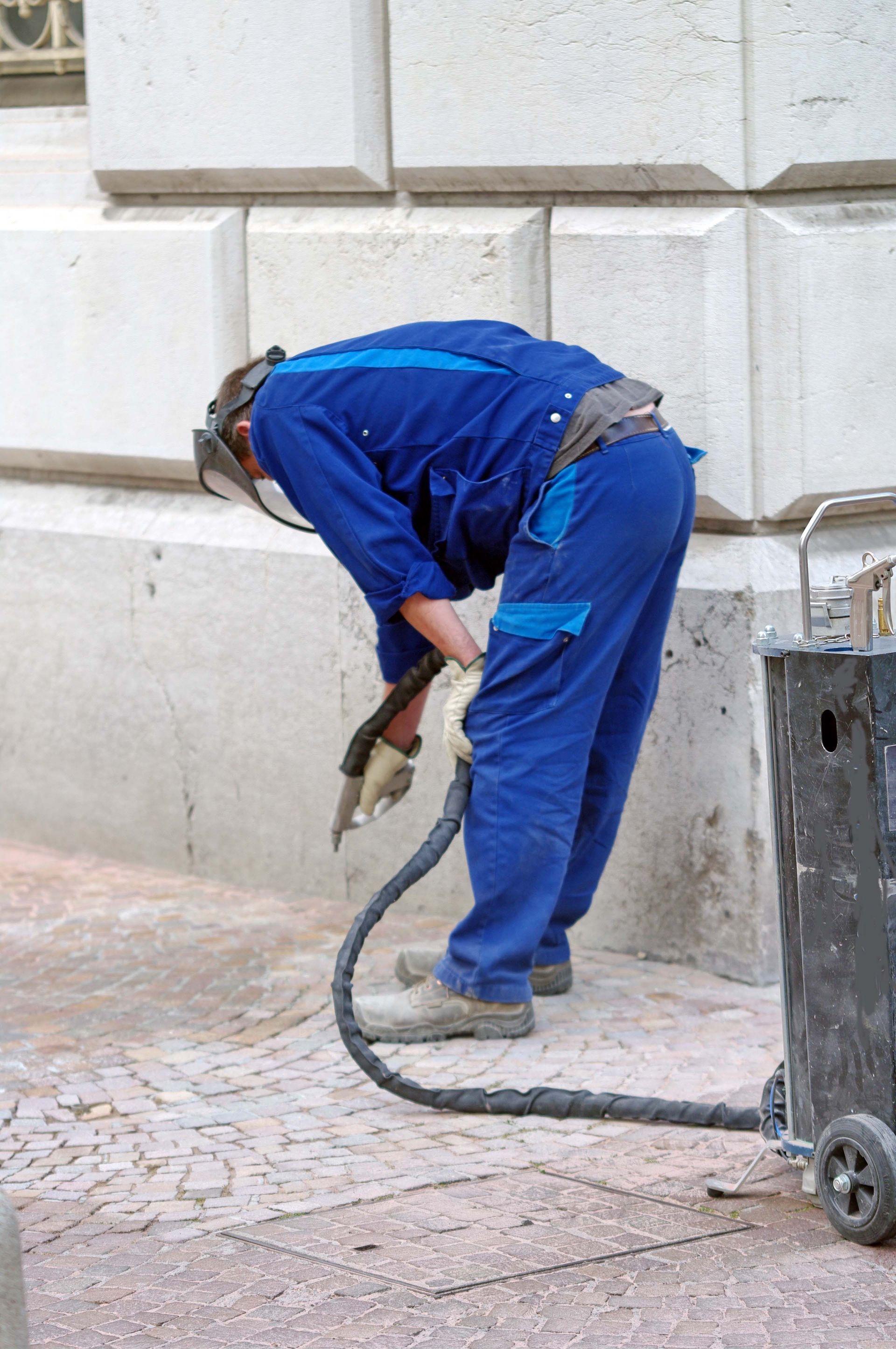 Un ouvrier en salopette bleu sable les pavés d'une rue à l'aide d'un tuyau d'arrosage et portant un équipement de protection.