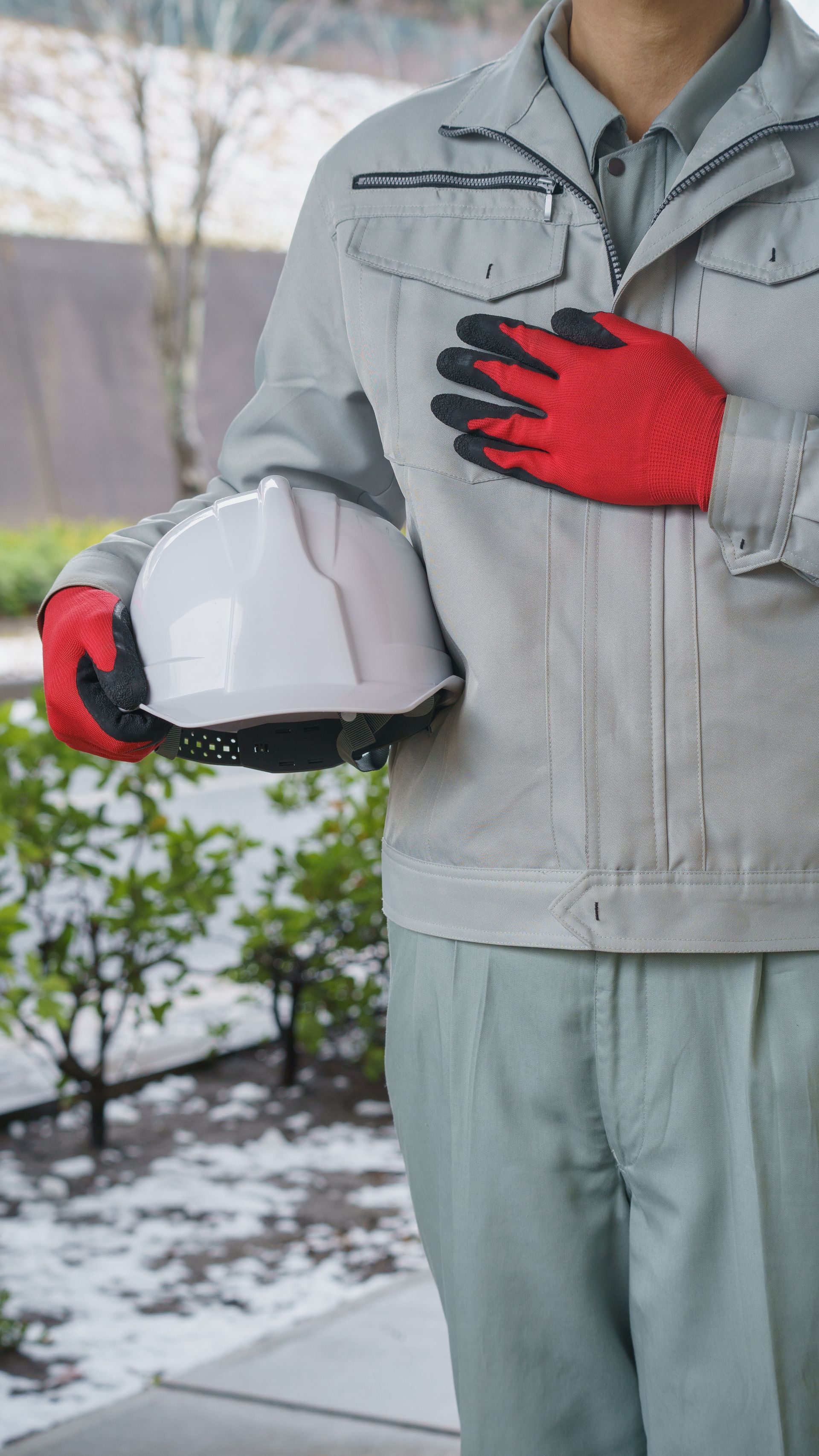 Ouvrier en uniforme gris, gants rouges, tenant un casque blanc, main sur la poitrine, à l'extérieur.