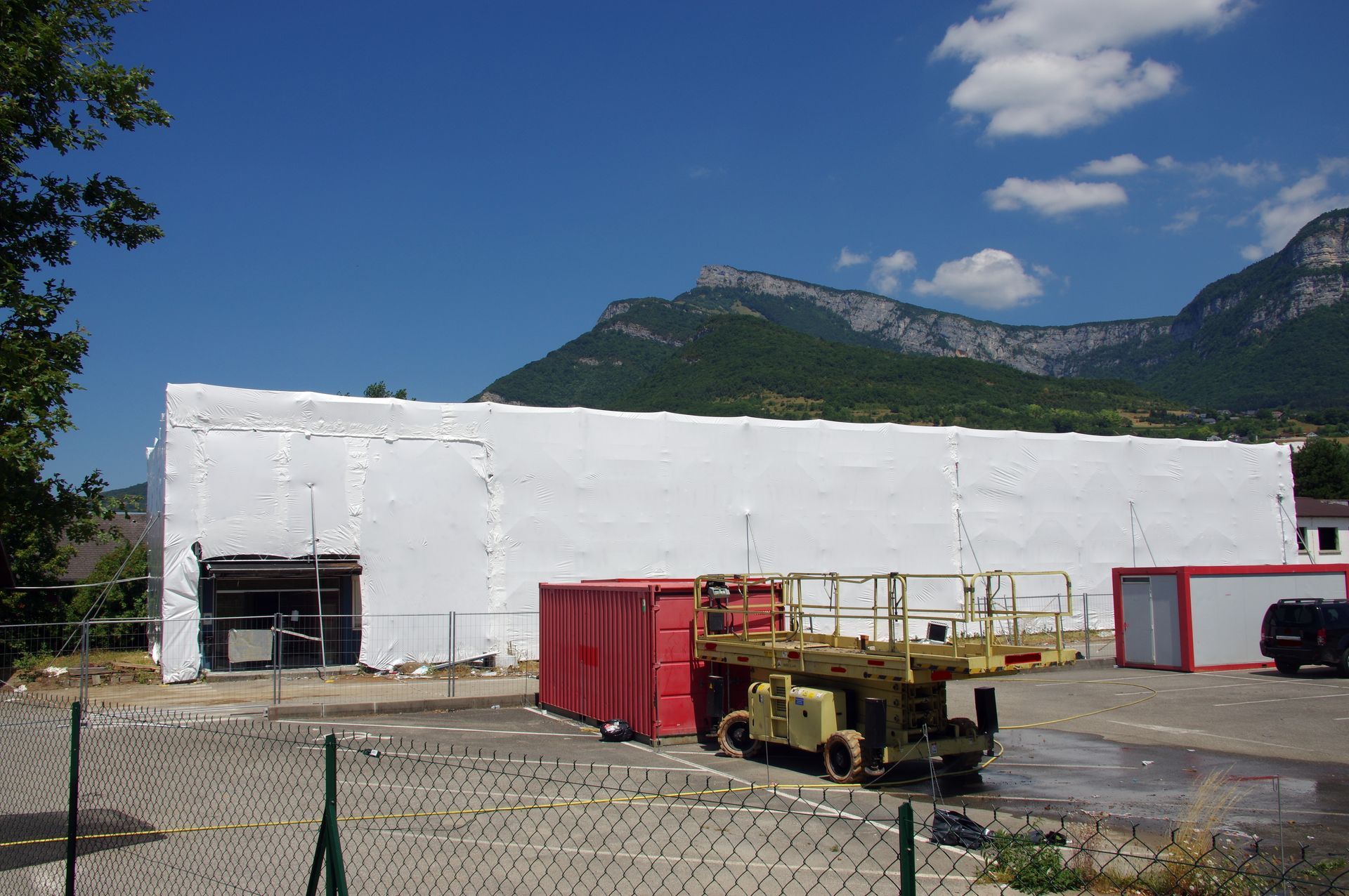 Extérieur du bâtiment avec revêtement blanc, conteneurs rouges, ascenseur jaune et montagnes en arrière-plan sous un ciel bleu.