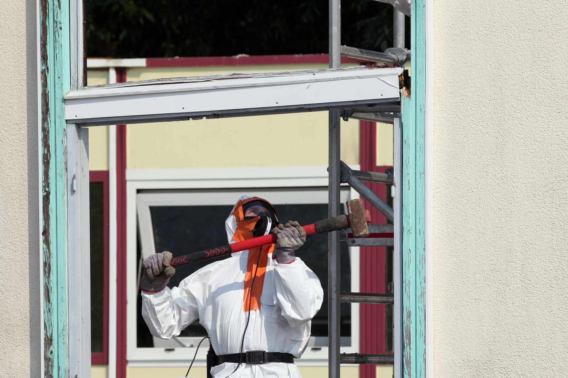 Une personne en tenue de protection démolit un cadre de fenêtre à coups de masse. Extérieur du bâtiment.