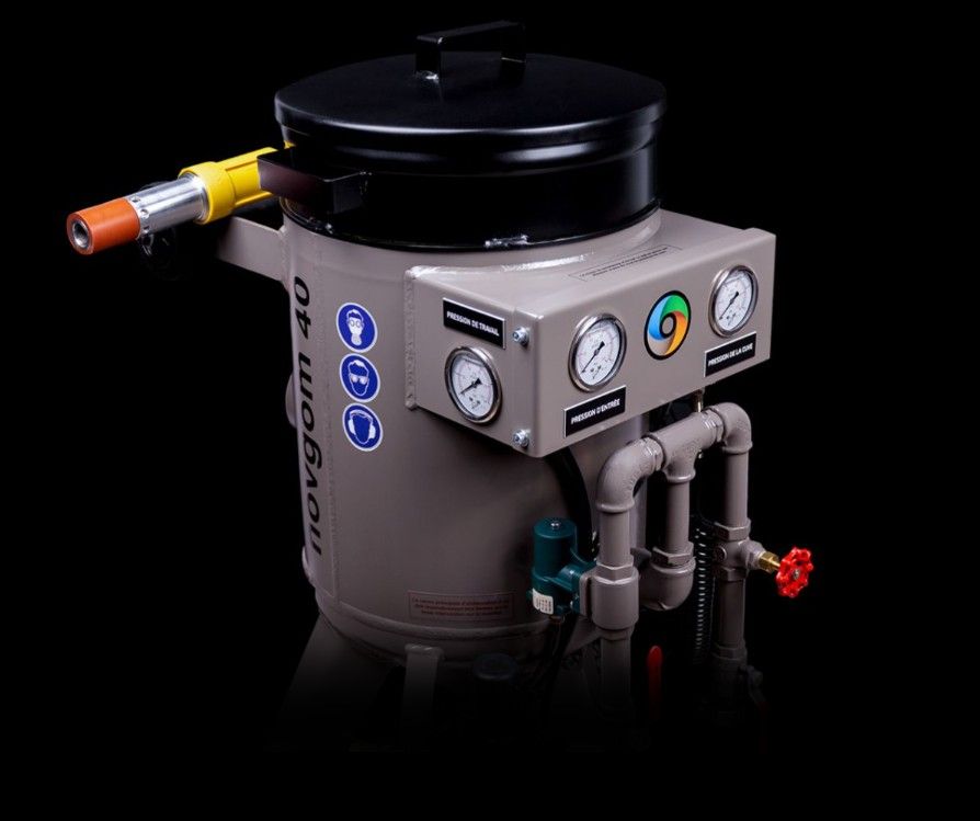 Un ouvrier en salopette bleu sable les pavés d'une rue à l'aide d'un tuyau d'arrosage et portant un équipement de protection.