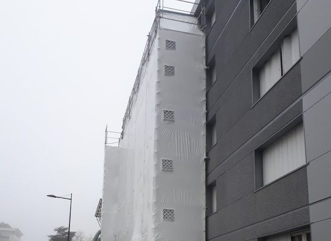 Bâtiment partiellement recouvert de bâche blanche à rénover, à côté d'un bâtiment gris.