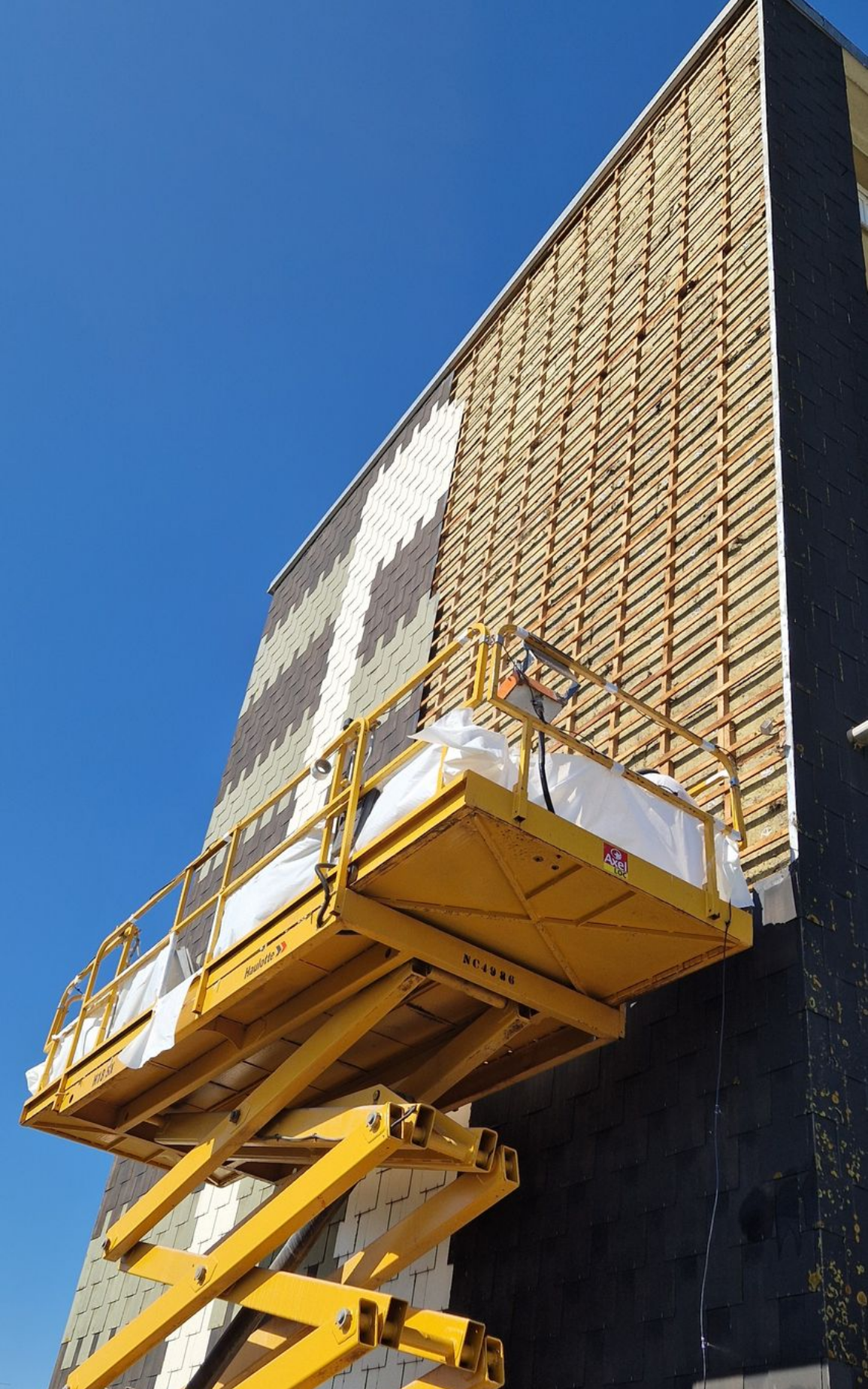 Plateforme d'ascenseur jaune près d'un bâtiment en construction à la façade texturée, sur fond de ciel bleu.