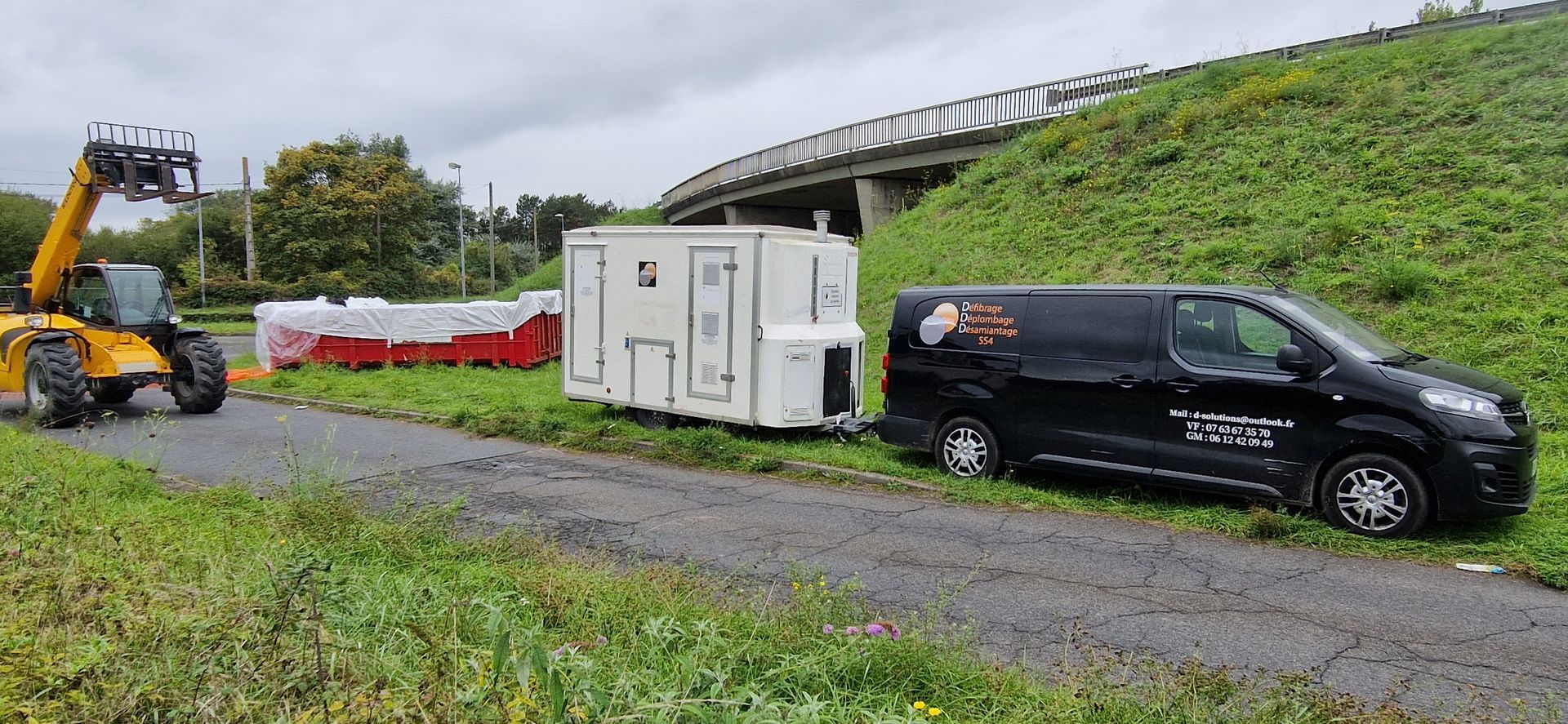 Une remorque blanche, des matériaux rouges et une camionnette noire en bord de route, près d'un pont. Intervention pour le retrait de déchets d'amiante.