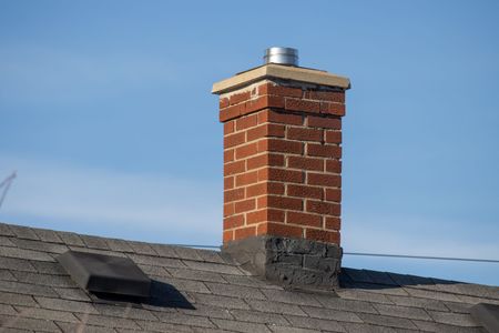 Une cheminée en briques coiffée d'un chapeau métallique se détache sur un toit de bardeaux, se détachant sur un ciel bleu limpide.
