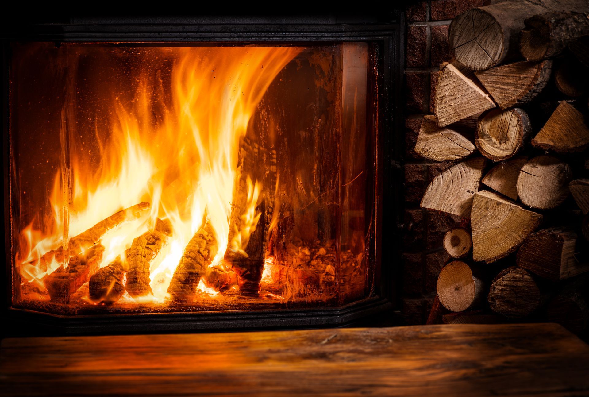 Des flammes d'un orange vif brûlent dans une cheminée vitrée, avec une pile de bûches de bois à proximité, sur une surface en bois.