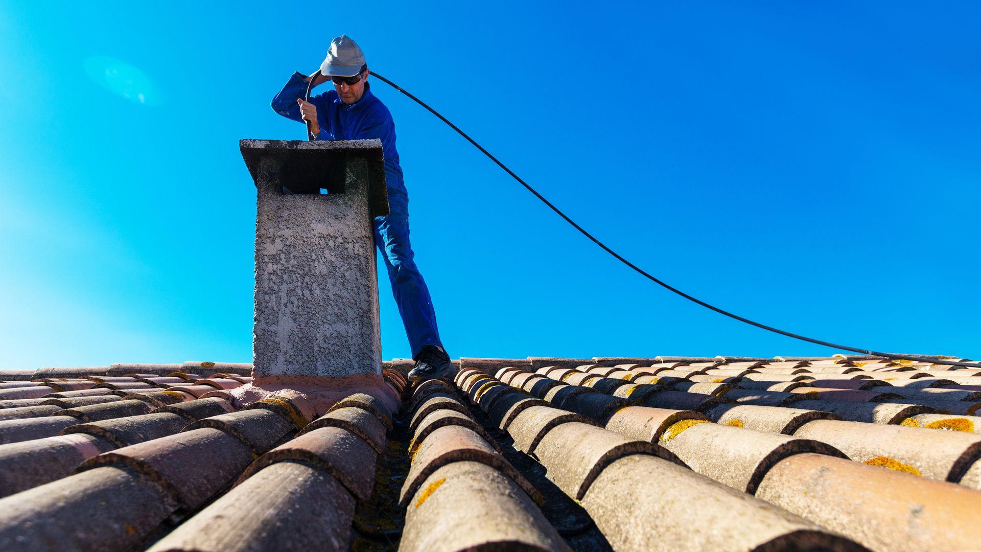 Une personne en tenue de travail et casque de chantier nettoie une cheminée sur un toit de tuiles, sur fond de ciel bleu éclatant.