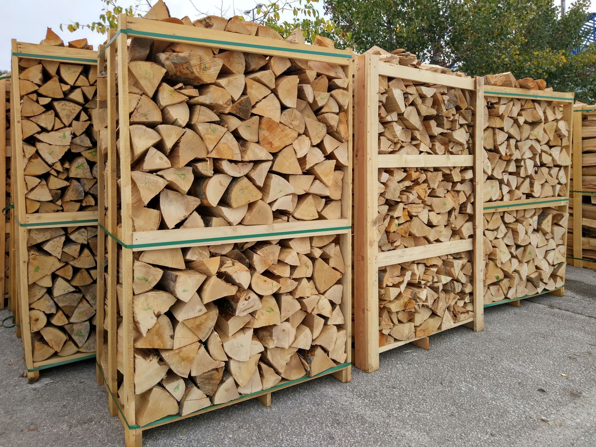 Plusieurs palettes en bois, sur lesquelles sont empilées des bûches fendues et soigneusement disposées, sont posées à l'extérieur sur un sol gravillonné.