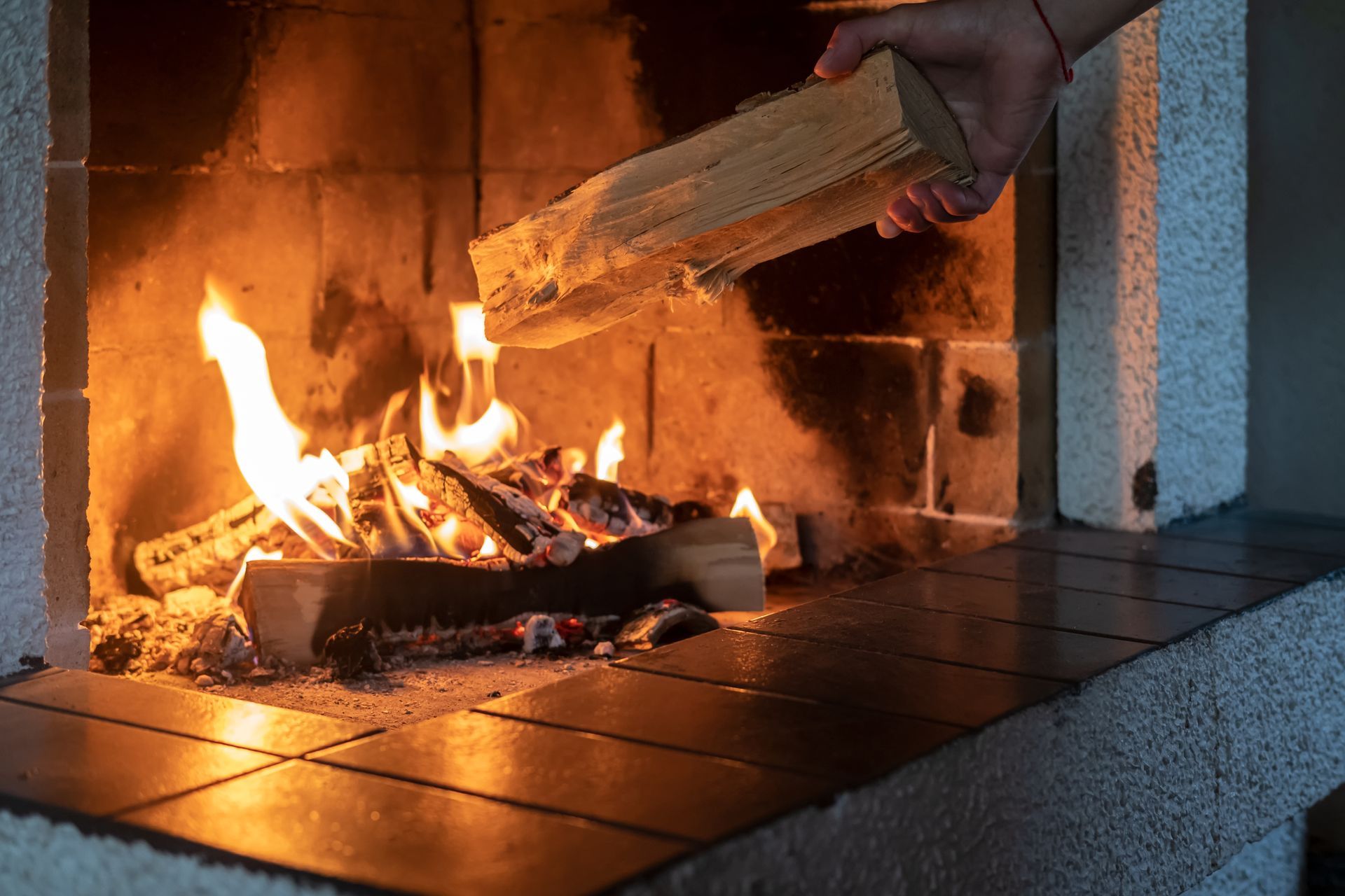 Une main dépose une bûche de bois dans une cheminée où brûlent des flammes.