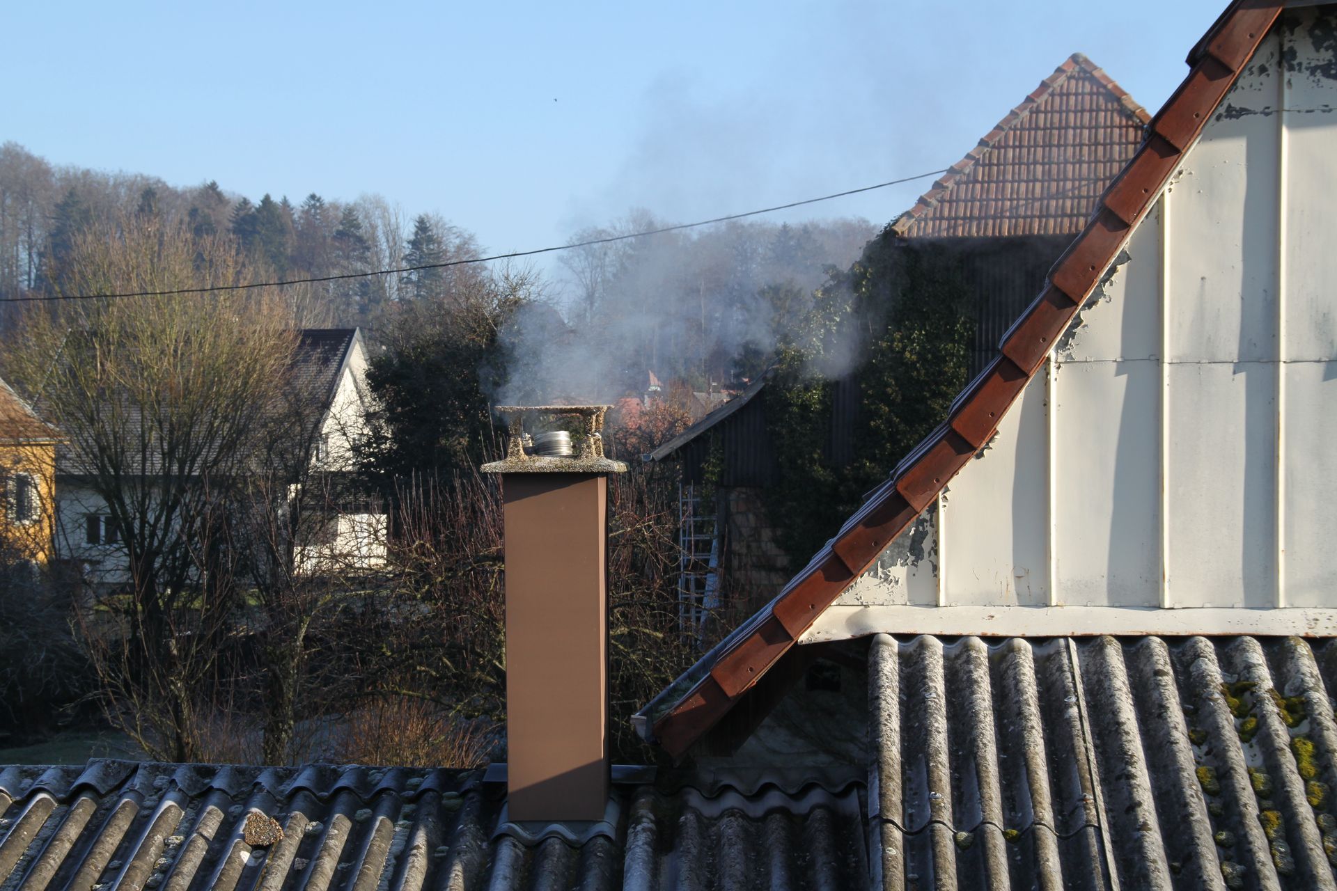 Une cheminée carrée et brune crache de la fumée sur fond de toit de tuiles, de bardage blanc et de flanc de colline par une journée ensoleillée.