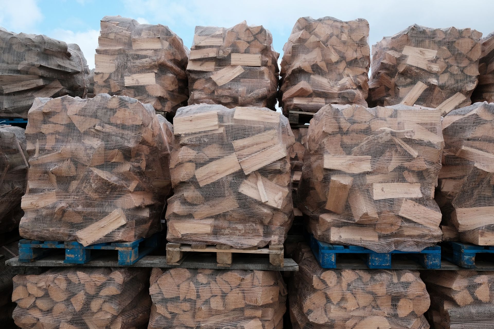 Des palettes de bûches de bois de chauffage, soigneusement empilées et enveloppées dans du filet, se détachent sur un ciel nuageux.
