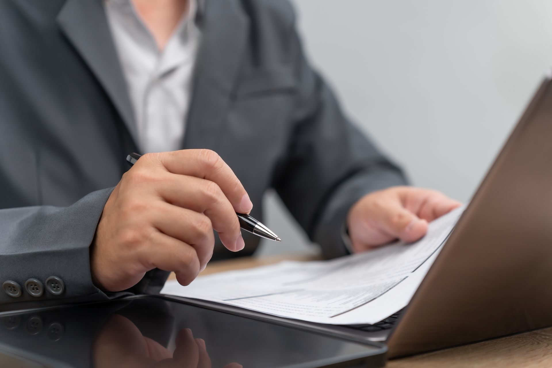 Eine Person in einer dunklen Jacke schreibt an einem Schreibtisch neben einem Laptop auf Papier.