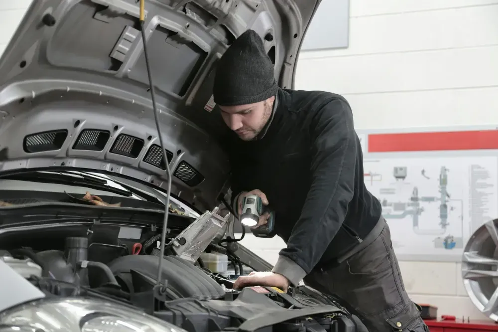 Un técnico con un suéter negro y un gorro de lana inspecciona con una linterna el motor abierto de un automóvil en un taller.