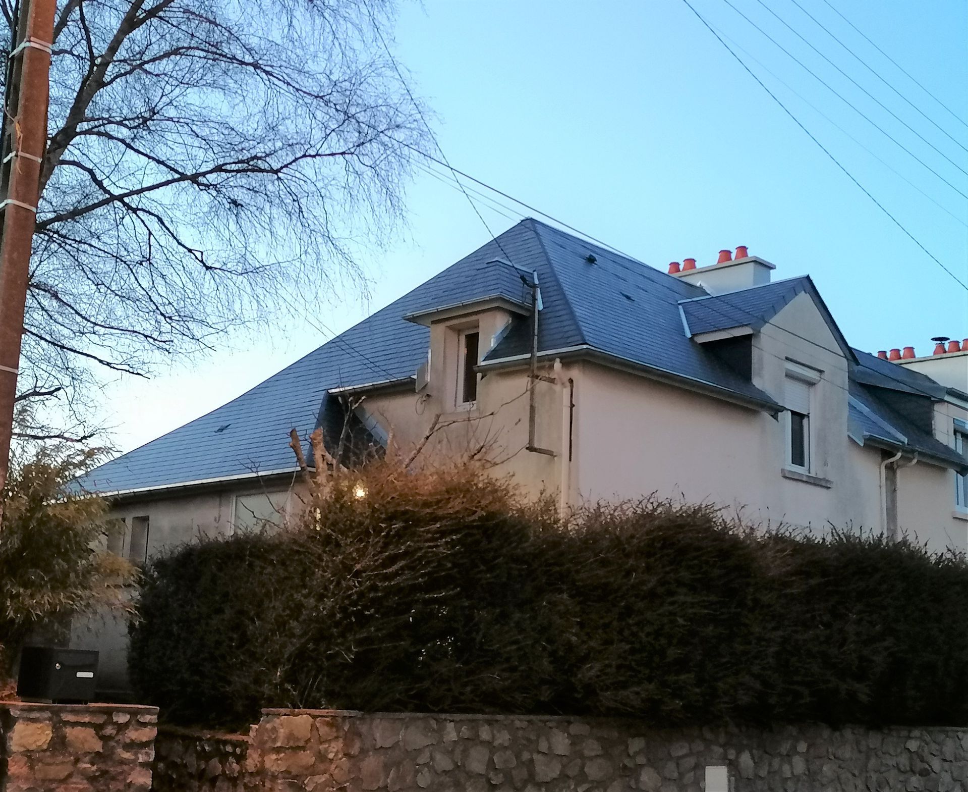 Une maison beige au toit d'ardoise, derrière de hautes haies et un mur de pierre