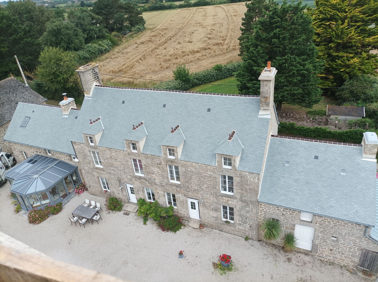 Vue aérienne d'une ferme en pierre à la campagne, avec des toits en ardoise, une terrasse et des champs à l'arrière.