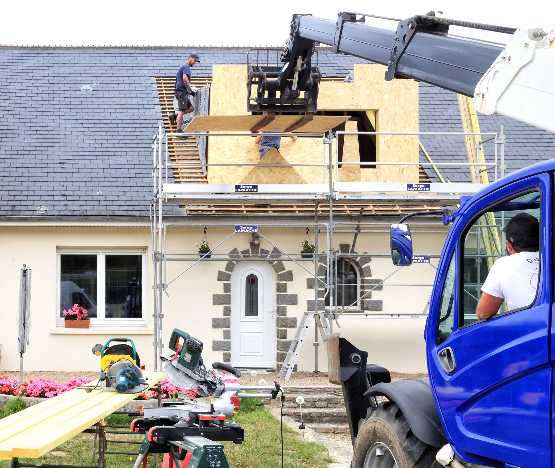 Des ouvriers réparent le toit d'une maison à côté d'un camion bleu équipé d'une grue