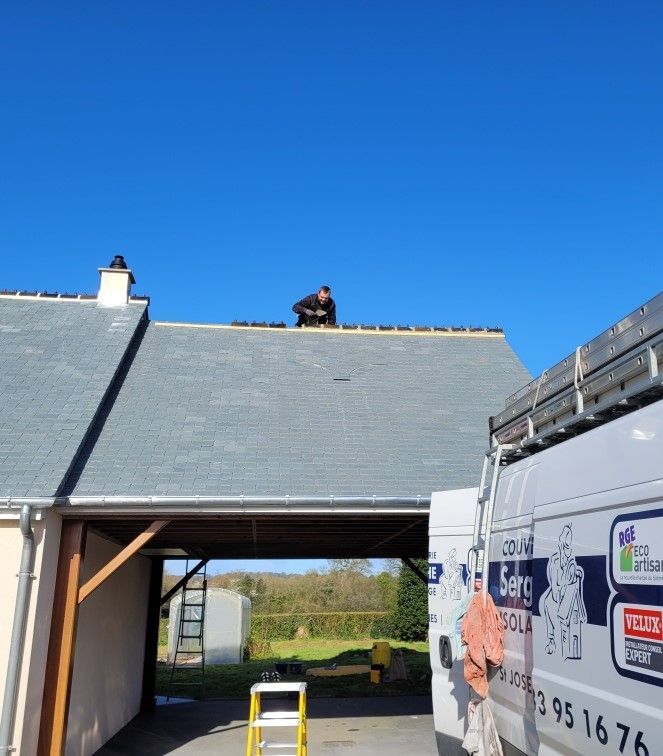 Maison en construction au toit d'ardoise, avec une camionnette à droite et un ciel bleu dégagé