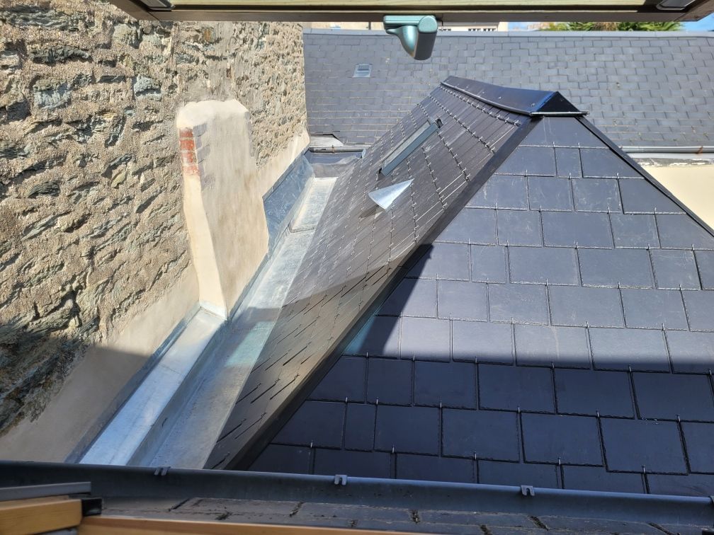 Toit en ardoise à forte pente avec une noue en zinc jouxtant un mur en pierre du côté gauche.