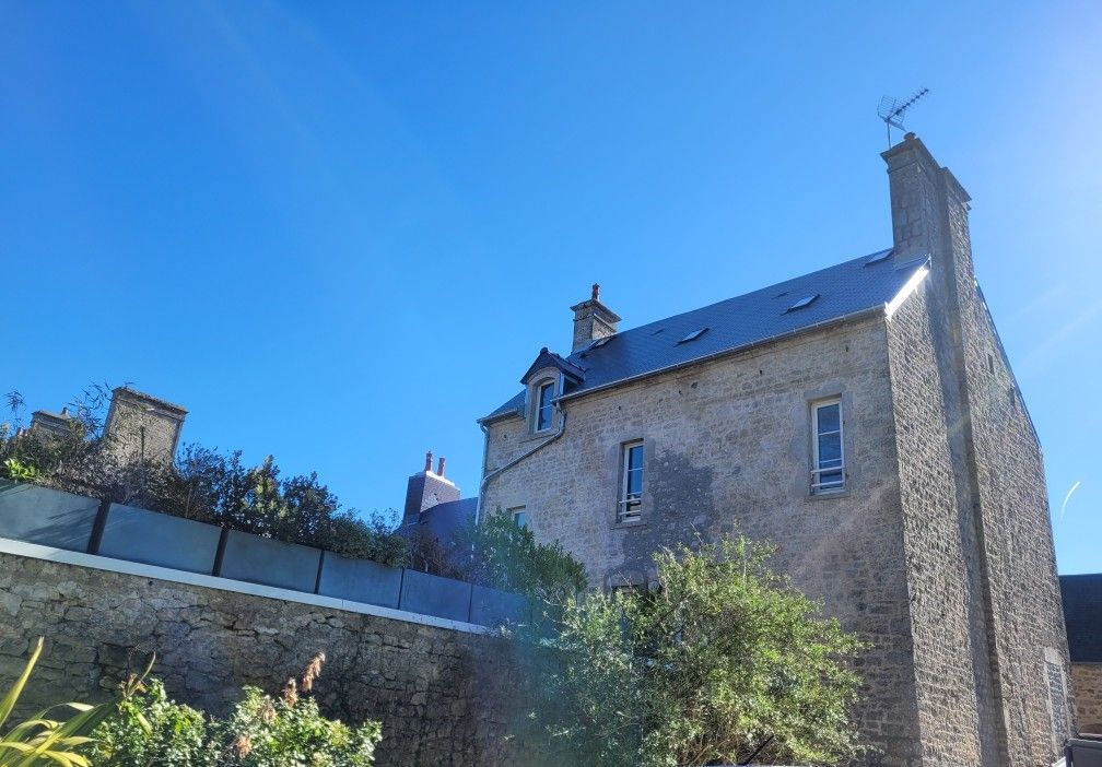 Maison en pierre sous un ciel bleu, avec des arbres et un muret au premier plan