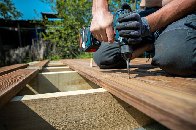 Une personne utilise une perceuse électrique pour fixer une planche de terrasse en bois à l'extérieur.