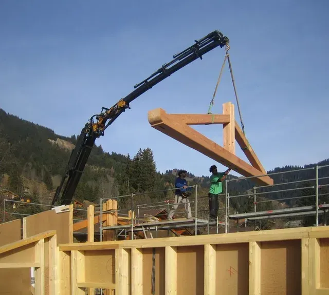 Une grue soulève une ferme de toit en bois sur un chantier de construction, deux ouvriers se trouvant en dessous.