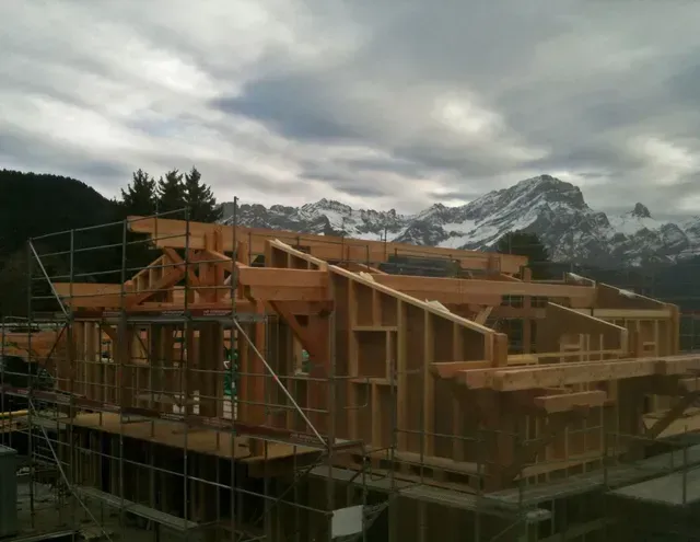 Maison à ossature bois en construction, avec des échafaudages, se détachant sur des montagnes nuageuses.