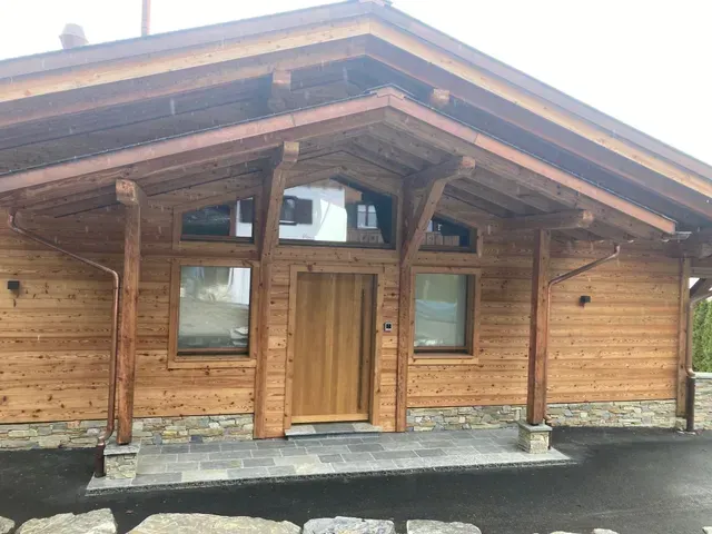 Extérieur d'un bâtiment de style cabane en bois avec porche, porte d'entrée et fenêtres sous un toit en pente.